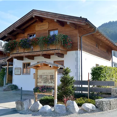 Ein traditionelles Holzhaus mit einem Balkon und blühenden Pflanzen. Im Hintergrund sind sanfte Hügel zu sehen.