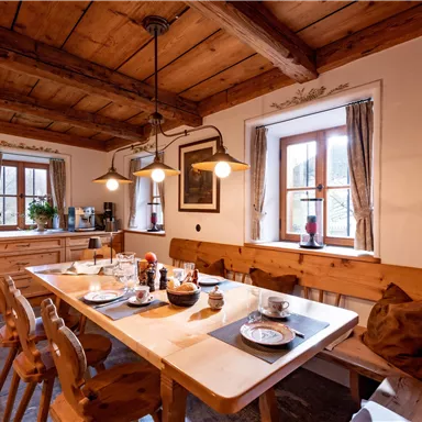 Eine gemütliche Küche mit Holzmöbeln und einem großen Tisch. Der Tisch ist gedeckt und das Licht fällt sanft durch die Fenster.