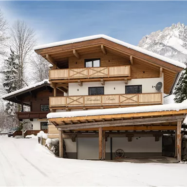 Ein traditionelles Chalet im Winter mit Holzverkleidung und einem schneebedeckten Dach. Im Hintergrund sind verschneite Bäume und Berge zu sehen.