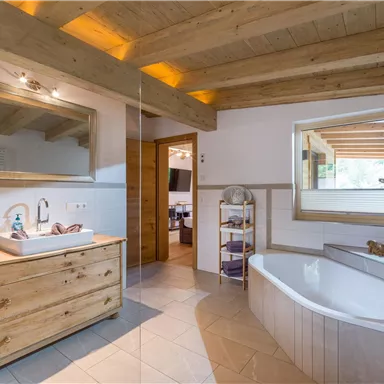 Ein modernes Badezimmer mit Holzbalkendecke und eleganter Ausstattung. Es gibt eine Badewanne, ein Waschbecken und große Fenster, die viel Licht hereinlassen.