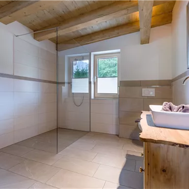 Ein modernes Badezimmer mit einer geräumigen Dusche und Holzdetails. Helle Fliesen und Fenster sorgen für viel Licht.