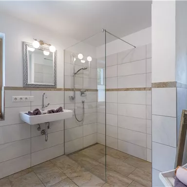 Ein modernes Badezimmer mit einer Glasdusche und einem weißen Waschbecken. Die Wände sind hell gefliest und es gibt natürliche Lichtquelle durch ein Fenster.