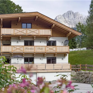 Ein schönes Holzhaus in den Bergen mit einem Balkon und umgeben von grüner Natur. Im Hintergrund sind beeindruckende Berge zu sehen.