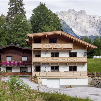 Ein gemütliches zweistöckiges Haus im alpinen Stil, umgeben von grünen Bäumen. Im Hintergrund erheben sich majestätische Berge.