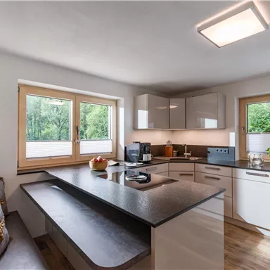 Eine moderne Küche mit hellen Schränken und einer eleganten Frühstücksbar. Große Fenster sorgen für viel natürliches Licht und Blick ins Freie.