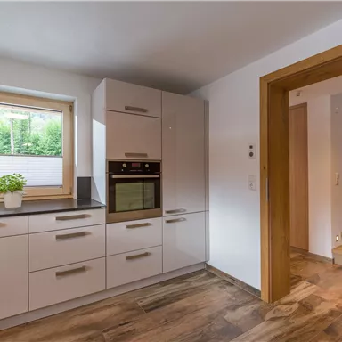 Eine moderne Küche mit hellen Schränken und einem Backofen. Im Hintergrund sind eine Treppe und ein Fenster mit Blick ins Grüne zu sehen.