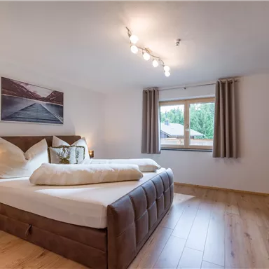 Ein modernes Schlafzimmer mit einem großen Bett und bequemen Kissen. Große Fenster sorgen für viel Tageslicht und einen Blick ins Grüne.