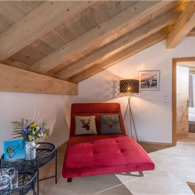 Ein gemütlicher Raum mit einer roten Liege und einem modernen Nachttisch. Die Holzbalkendecke und die warmen Farben schaffen eine einladende Atmosphäre.