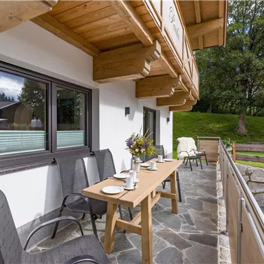 Ein einladender Balkon mit Holzverkleidung und einem Tisch, gedeckt für eine Mahlzeit. Im Hintergrund sind Grünflächen und Bäume sichtbar.