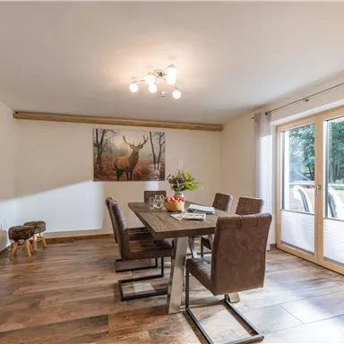Ein modernes Esszimmer mit einem großen Holztisch und bequemen Stühlen. Ein Bild eines Rehs hängt an der Wand und große Fenster lassen viel Licht herein.