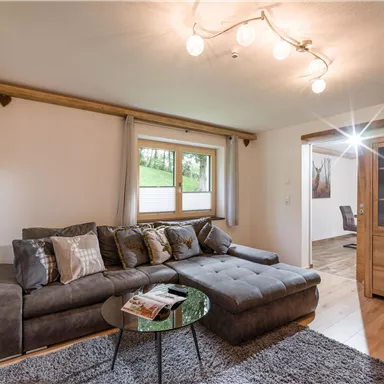 Ein gemütliches Wohnzimmer mit einem grauen Sofa und einem runden Tisch. Helle Wände und große Fenster sorgen für eine freundliche Atmosphäre.