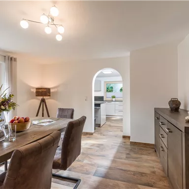 Ein einladendes Esszimmer mit einem Holztisch, Stühlen und einer modernen Deckenleuchte. Im Hintergrund ist eine offene Küche und ein großes Fenster, das viel Tageslicht hereinlässt.