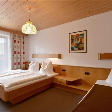 Ein helles Hotelzimmer mit zwei Betten und Holzdecke. Es gibt ein großes Fenster mit Vorhängen und ein Kunstwerk an der Wand.