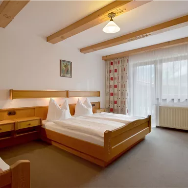 Ein gemütliches Schlafzimmer mit zwei Betten und Holzmöbeln. Große Fenster lassen viel Licht herein und der Raum hat eine helle, freundliche Atmosphäre.