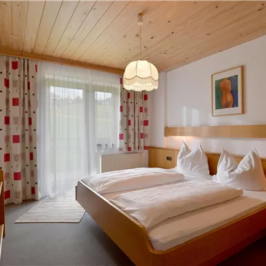 Ein gemütliches Schlafzimmer mit zwei Betten, Holzdecke und einem Hängelicht. Große Fenster mit Vorhängen und ein modernes Kunstwerk an der Wand.