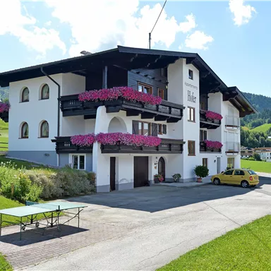 Ein ruhiges Landhaus mit Balkonen voller blühender Blumen. Im Vordergrund gibt es einen Tisch und eine gelbe Auto auf der gepflasterten Einfahrt.
