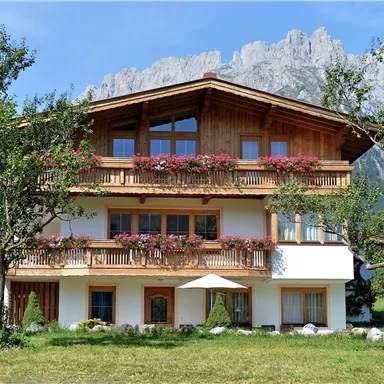 Ein charmantes alpenländisches Haus mit Balkonen und Blumenschmuck. Im Hintergrund sind beeindruckende Berge zu sehen.