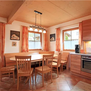 Eine moderne Küche mit einem großen Esstisch und Holzstühlen. Das Licht fällt durch die Fenster und schafft eine einladende Atmosphäre.