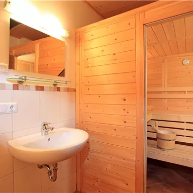 Ein helles Badezimmer mit einem Waschbecken und einem großen Spiegel. Neben dem Waschbecken befindet sich eine Sauna mit Holzverkleidung.