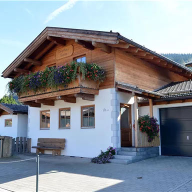 Ein schönes Holzhaus mit einem Balkon voller Blumen. Der klare Himmel und die Berge im Hintergrund verleihen dem Bild eine idyllische Atmosphäre.
