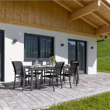 Eine moderne Terrasse mit einem Esstisch und Stühlen. Im Hintergrund ist ein grüner Hang und große Fenster sichtbar.