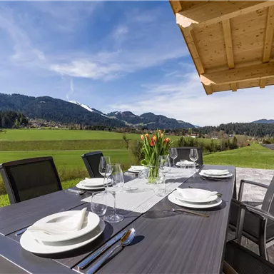 Ein eleganter Essbereich im Freien mit Blick auf grüne Wiesen und Berge. Der Tisch ist schön gedeckt mit Geschirr und Blumen.
