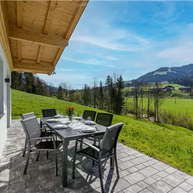 Eine moderne Terrasse mit einem Esstisch und Stühlen. Im Hintergrund erstrecken sich grüne Wiesen und Berge unter einem klaren blauen Himmel.
