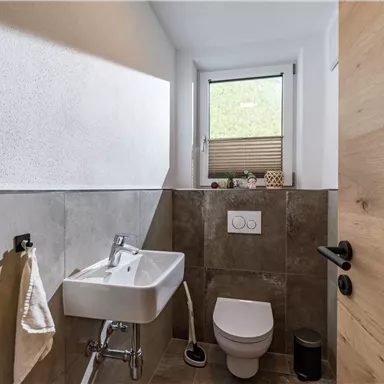 Ein modernes Badezimmer mit grauen Fliesen und einem kleinen Fenster. Es gibt ein Waschbecken und eine Toilette, sowie natürliche Holz-Elemente.