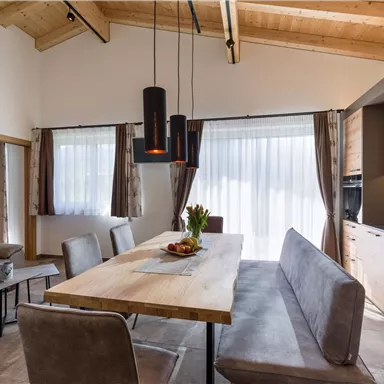 Ein modernes Esszimmer mit einem großen Holztisch und bequemen Sitzbänken. Die großen Fenster lassen viel Tageslicht herein und die Holzdecke sorgt für eine gemütliche Atmosphäre.