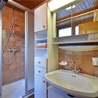 Ein modernes Badezimmer mit einer Dusche und einem Waschbecken. Die Wände sind in sanften Erdtönen gefliest, und es gibt genügend Stauraum.