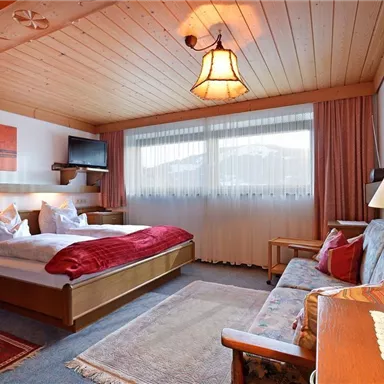 Ein gemütliches Hotelzimmer mit Holzmöbeln und einem großen Fenster, das viel Tageslicht hereinlässt. Es gibt ein Doppelbett, eine Sitzecke und eine warme, einladende Atmosphäre.