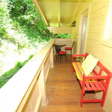 Ein gemütlicher Balkon mit roten Sitzgelegenheiten und einem kleinen Tisch. Im Hintergrund sieht man üppiges, grünes Laub.