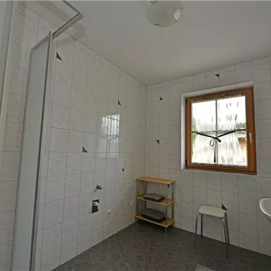 A bright bathroom with white tiles and a window. On the right side, there is a sink and a shower.