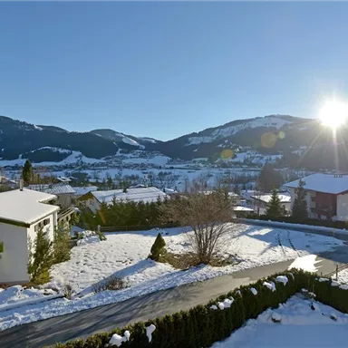 Eine winterliche Landschaft mit schneebedeckten Hügeln und Bergen. Die Sonne scheint über dem ruhigen Dorf mit Häusern und Bäumen.
