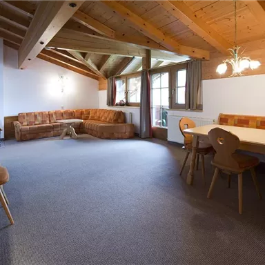 A cozy living space with wooden ceilings and a large window. A sofa, a dining table, and a television are also visible.