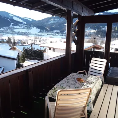 Ein Balkon mit einem Tisch und Stühlen, umgeben von schneebedeckten Bergen. Die Aussicht reicht über ein verschneites Tal und gemütliche Häuser.