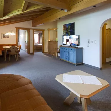 A cozy living room with a wooden ceiling and charming furniture. In the background, a television and a dining table can be seen.