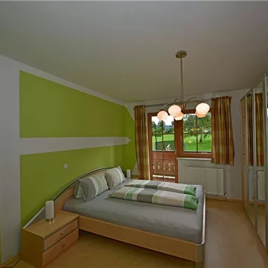 A modern bedroom with green walls and a large window. The room features a double bed and a wardrobe.