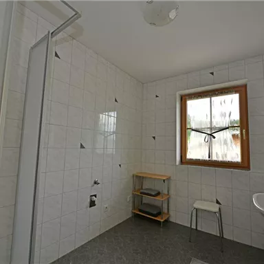 A simple bathroom with white tiles and a window. There is a shower, a sink, and a small shelf.