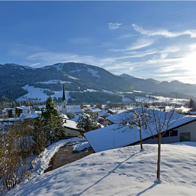 A picturesque winter landscape with snow-covered houses and mountains in the background. The sun shines brightly over the snowy scene.