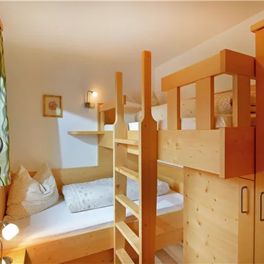 A cozy bedroom with a wooden loft bed and a single bed. The room has light walls and large windows that provide plenty of daylight.