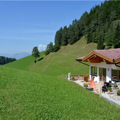 Ein wunderschönes Haus in der Natur, umgeben von saftigem Grün und sanften Hügeln. Im Hintergrund sind Wälder und ein klarer blauer Himmel sichtbar.