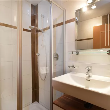 Ein modernes Badezimmer mit einer Dusche und einem Waschbecken. Die Wände sind hell gefliest und es gibt einen Spiegel über dem Waschbecken.