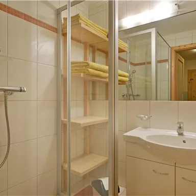 Ein modernes Badezimmer mit einer Dusche, einem Waschbecken und Regal für Handtücher. Die Wände sind gefliest und der Raum wirkt hell und einladend.