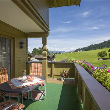 A sunny balcony with a table and chairs, surrounded by colorful flowers. The view stretches over green meadows and gentle hills into the distance.