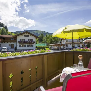 A cozy balcony with a red chair and a yellow parasol. In the background, beautiful mountains and traditional Austrian houses can be seen.