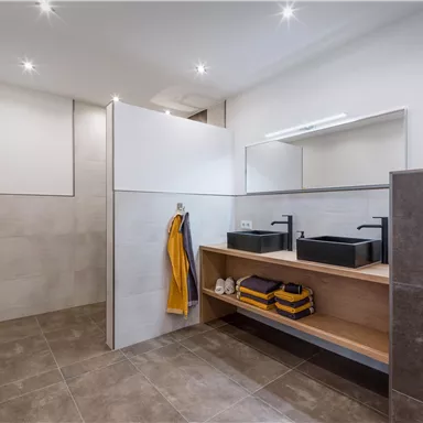 Ein modernes Badezimmer mit zwei schwarzen Waschbecken und Holzregalen. Helle Wände und stilvolle Beleuchtung schaffen eine einladende Atmosphäre.