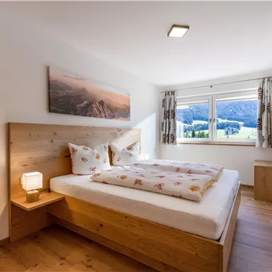Ein gemütliches Schlafzimmer mit einem Holzbett und einem großen Fenster. Der Raum ist hell und modern gestaltet, mit Blick auf die Berge.