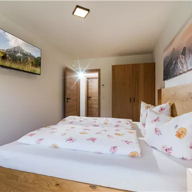 Ein gemütliches Schlafzimmer mit einem großen Bett und blühenden Kissen. An der Wand hängt ein Fernseher, und es gibt eine schöne Wanddekoration mit Berglandschaft.