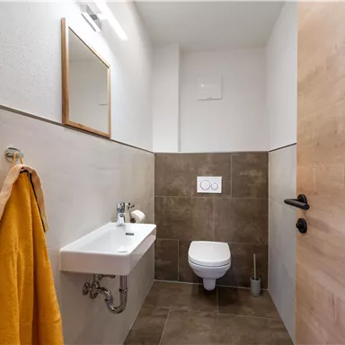 Ein modernes Badezimmer mit einem weißen Waschbecken und einer Toilette. An der Wand hängt ein orangefarbenes Handtuch, und es gibt einen Spiegel.